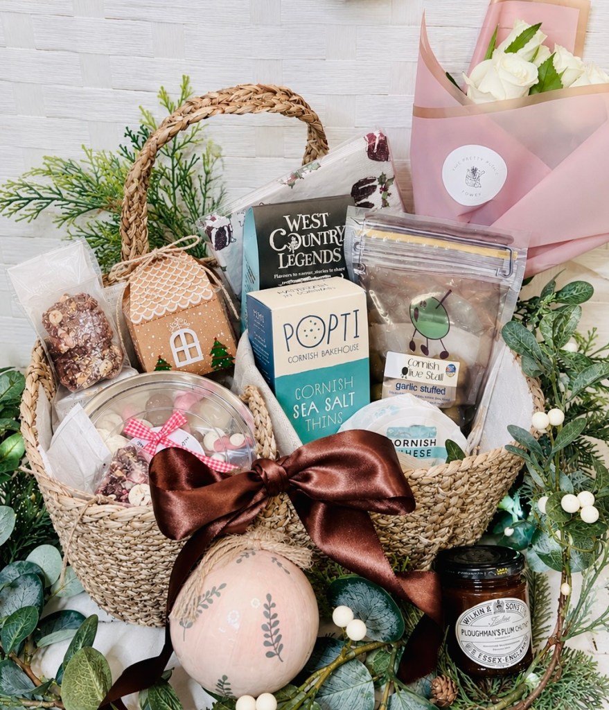 A beautifully arranged gift basket containing a variety of local delicacies, including Cornish sea salt thins, a gingerbread house, and assorted sweets, complemented by a bouquet of white roses and decorative greenery.