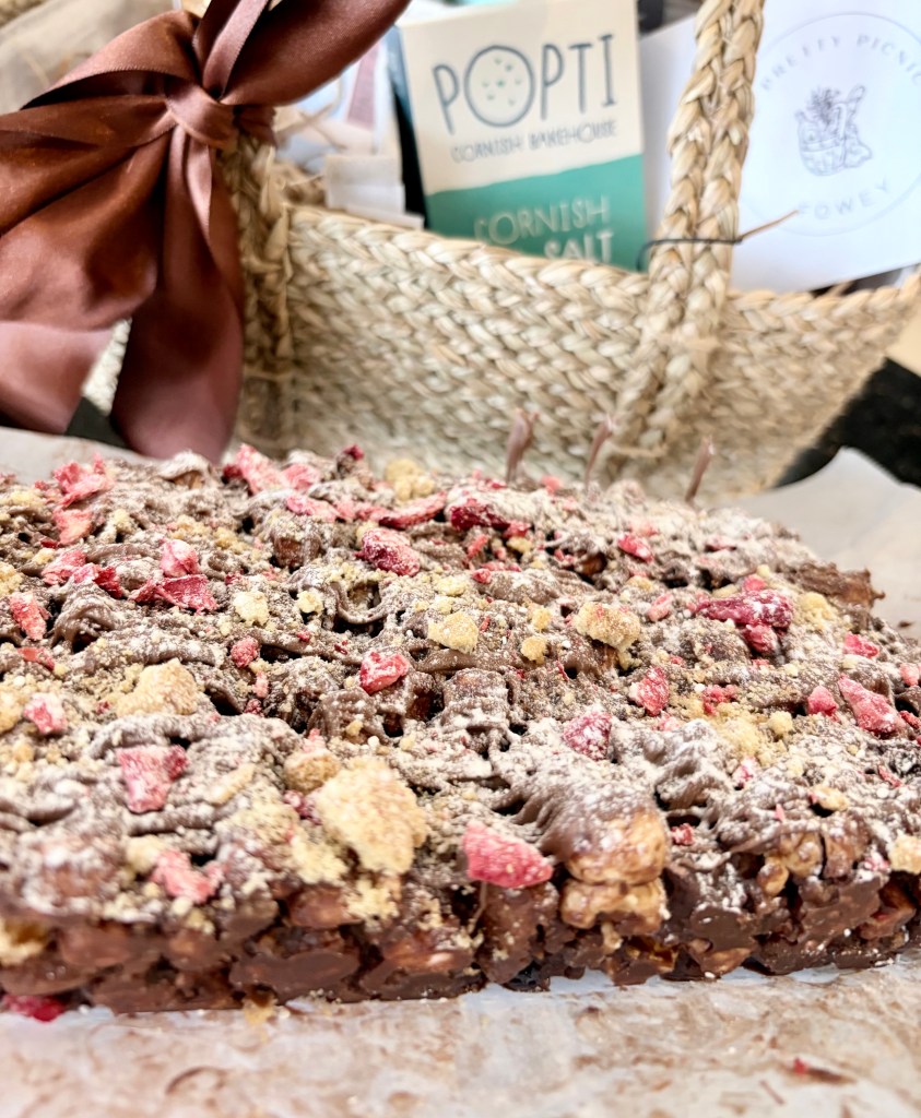 A close-up of a chocolate treat topped with crushed dried strawberries and biscuit crumbs, displayed on parchment paper with a woven basket in the background.