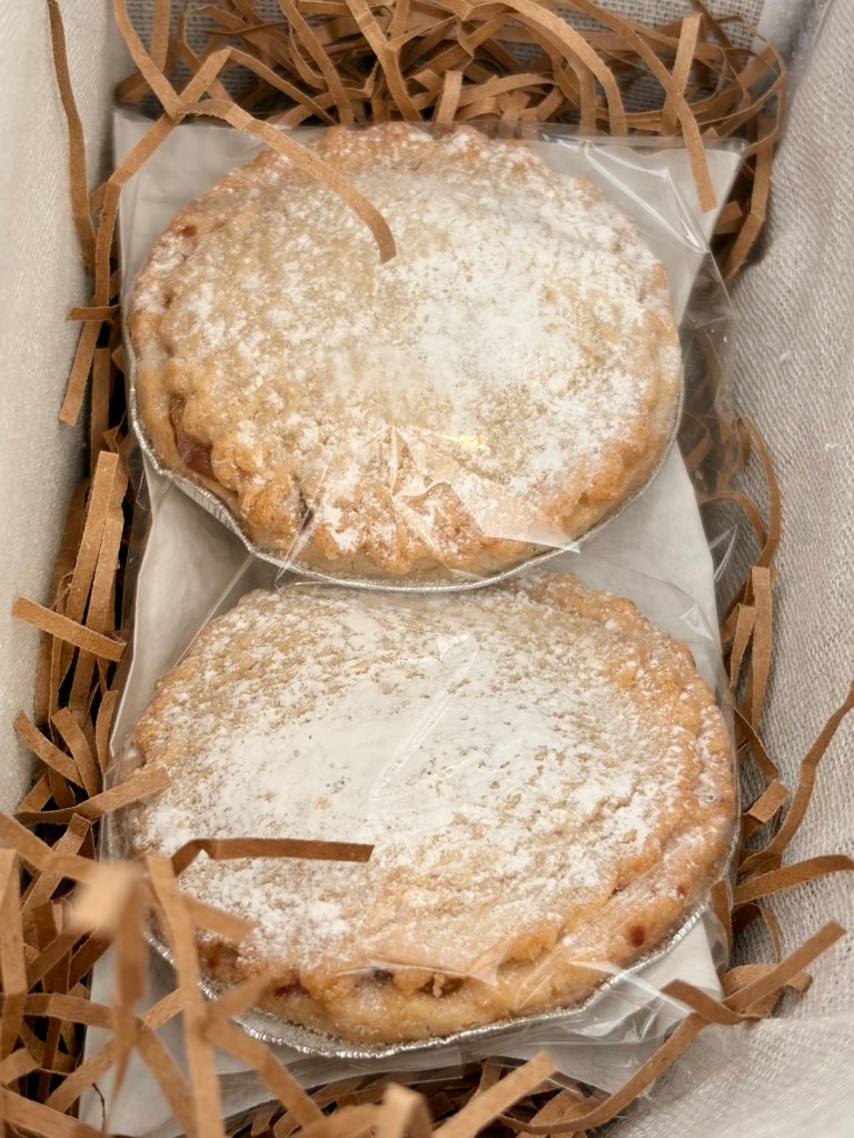 Two wrapped sweet pies dusted with icing sugar, resting in a bed of shredded paper.